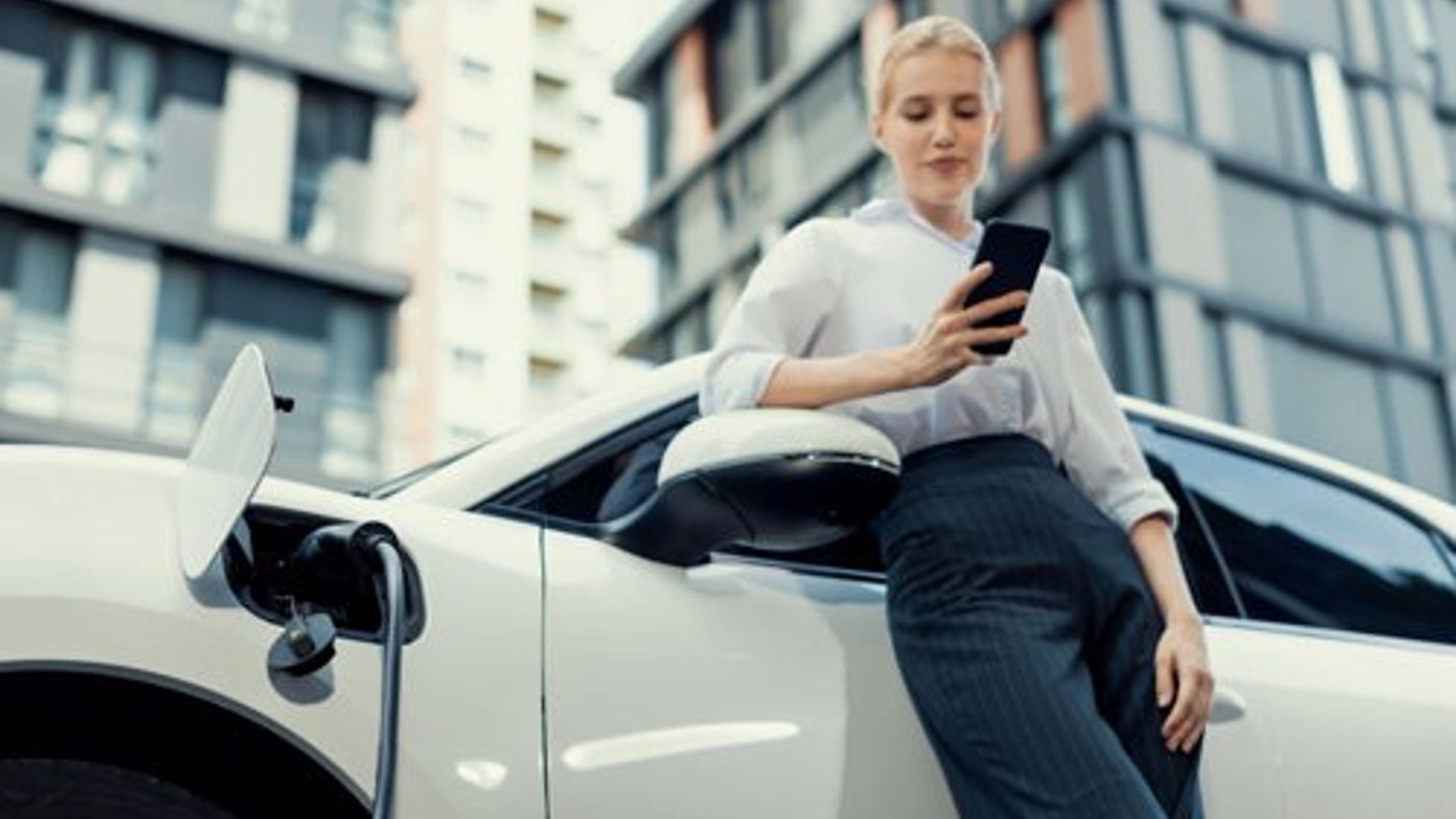 Woman leaning against a charging EV checking her phone.
