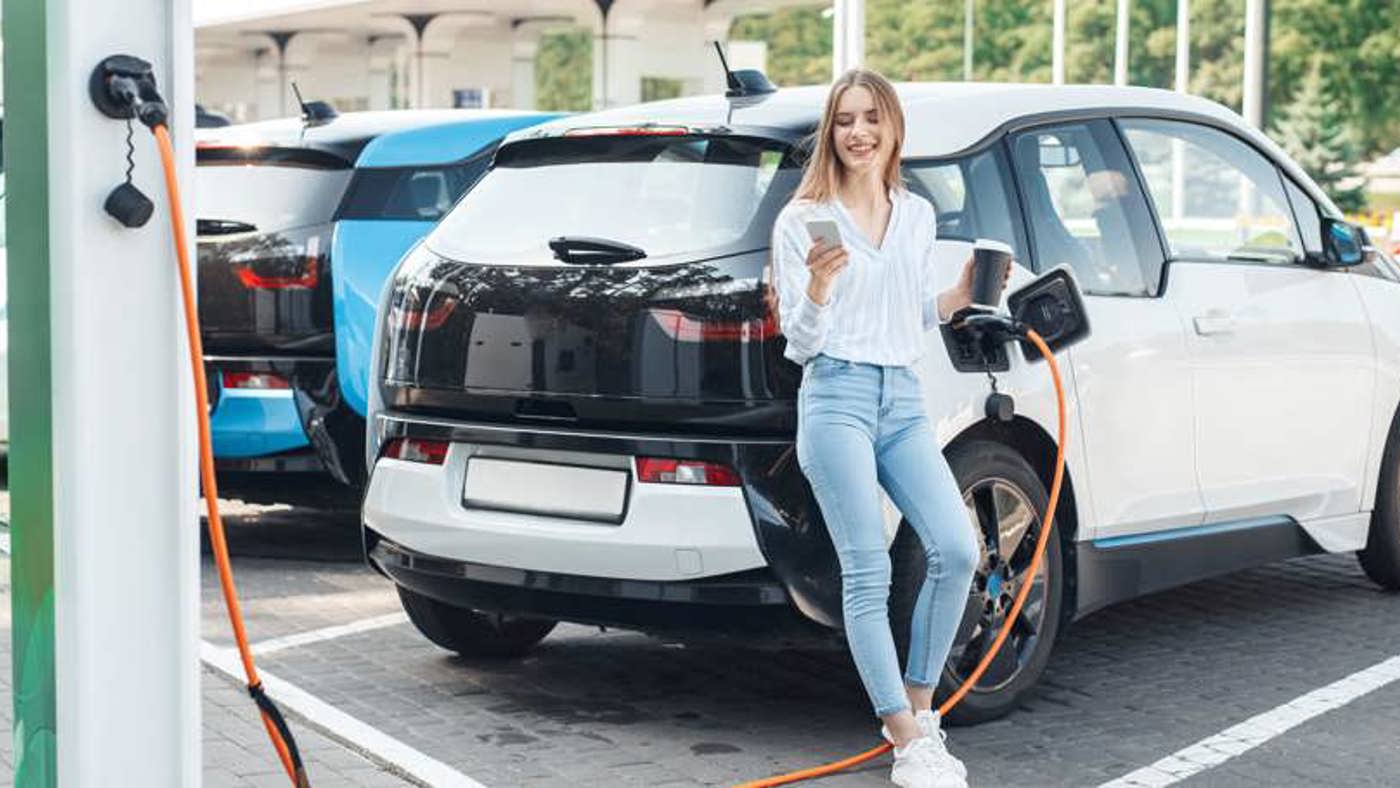 Woman leaning against her EV, smiling whilst checking her phone during a charging session.
